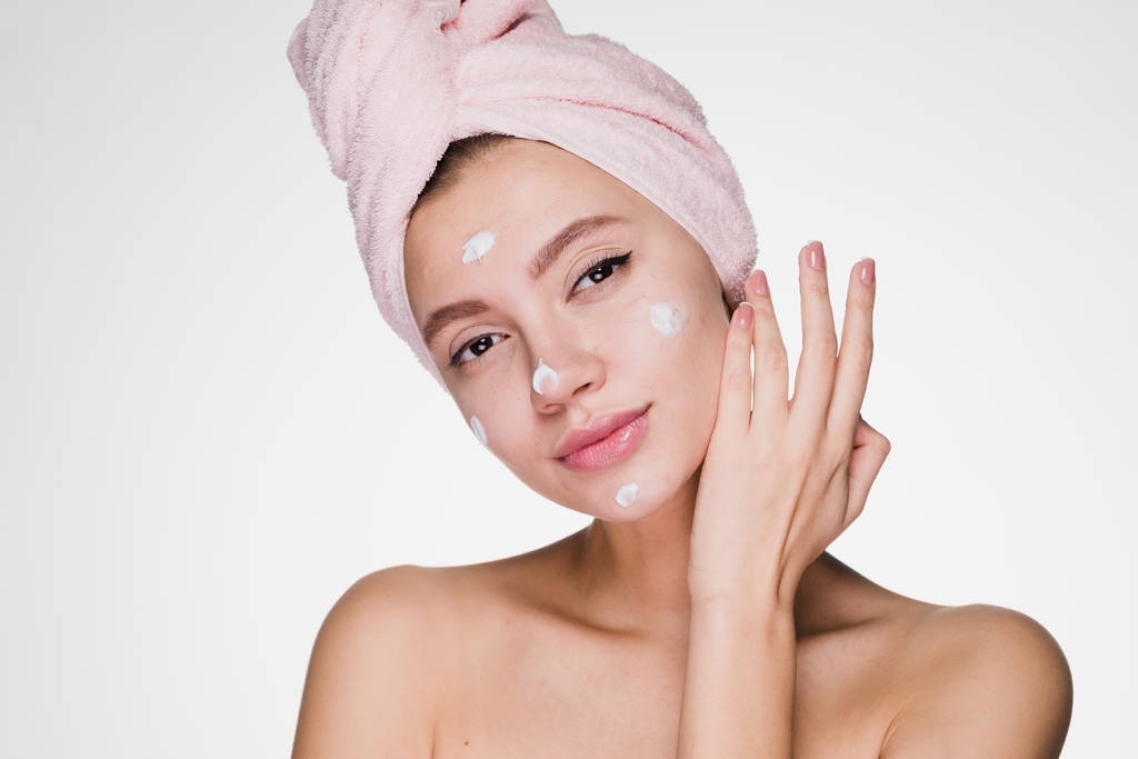 joven chica atractiva con toalla rosa en la cabeza aplica crema hidratante blanca en la cara
 - Foto, Imagen
