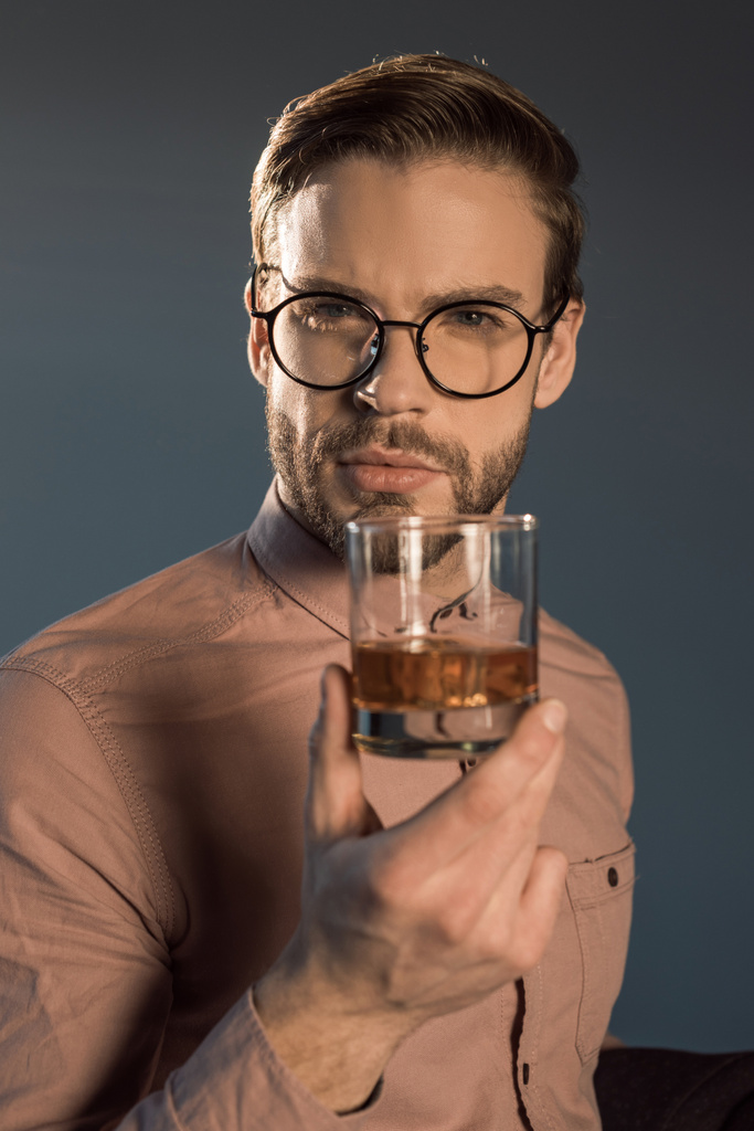 primo piano vista di elegante giovane uomo in occhiali in possesso di un bicchiere di whisky e guardando la fotocamera isolata su grigio
  - Foto, immagini