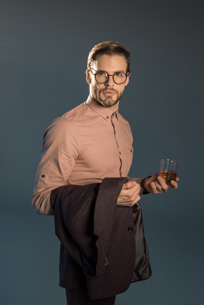 stylish young man in eyeglasses holding suit jacket and glass of whisky, looking at camera isolated on grey - Photo, Image