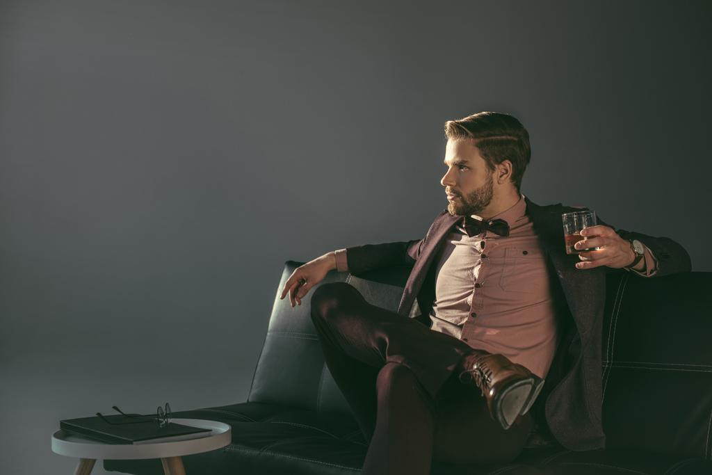 young businessman holding glass of whiskey and looking away isolated on grey - Photo, Image