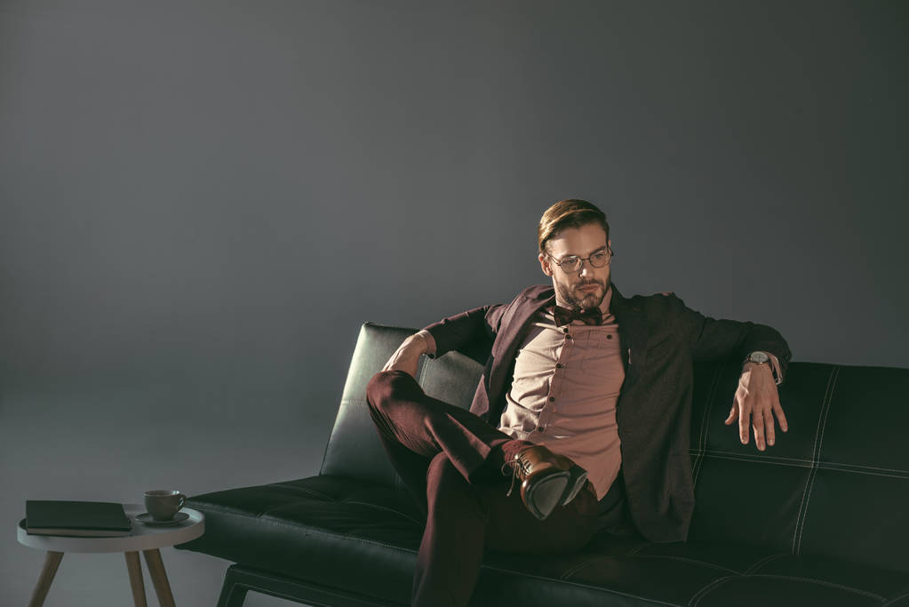 guapo joven hombre de negocios con estilo en gafas sentado en el sofá y mirando hacia otro lado en gris
  - Foto, Imagen