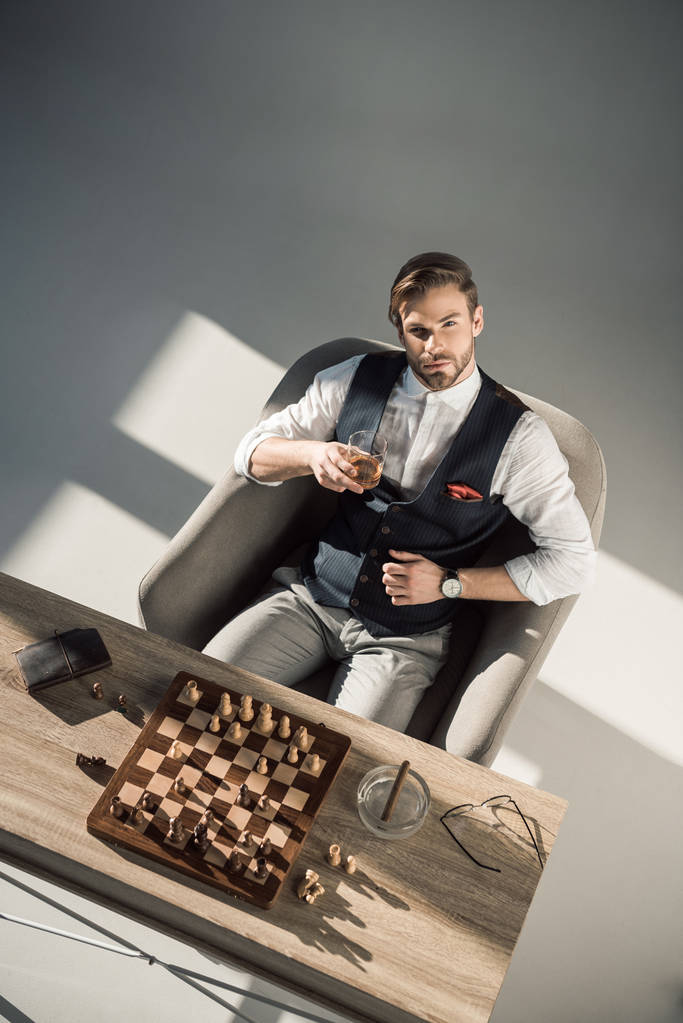 Aus der Vogelperspektive betrachtet blickt der junge Geschäftsmann in die Kamera, während er Whisky trinkt und Schach spielt  - Foto, Bild