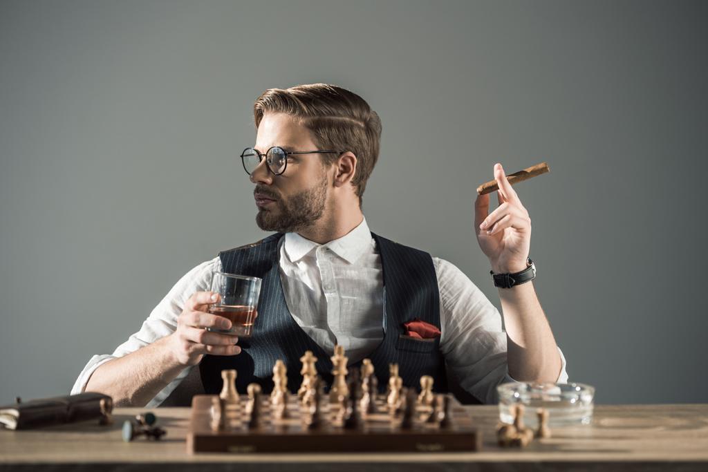young man with glass of whisky smoking cigar and looking away while playing chess  - Photo, Image