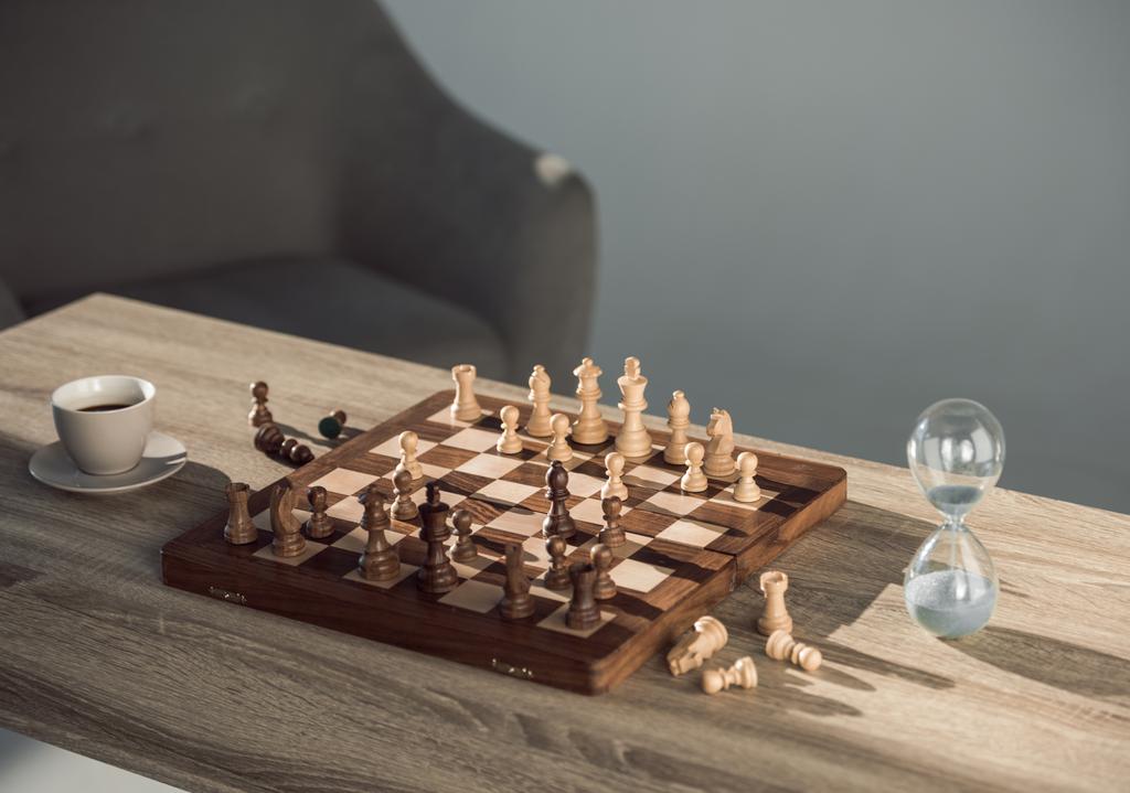 vergrote weergave van schaakbord met stukken, kopje koffie en zand klok op tafel   - Foto, afbeelding