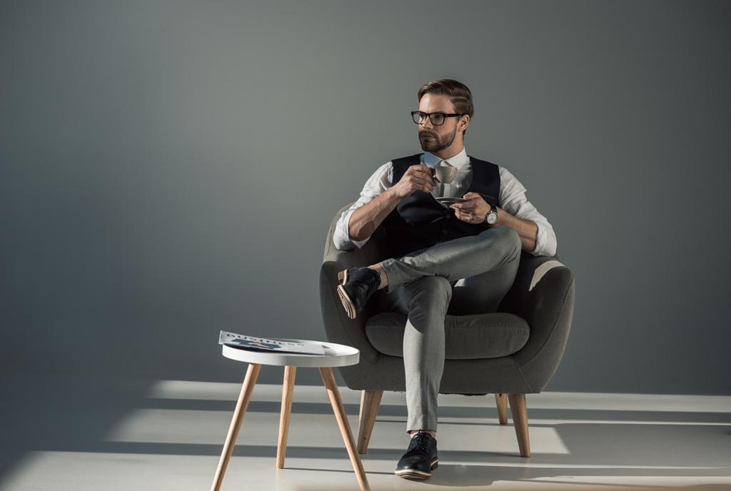 красивый стильный молодой человек сидит в кресле и смотрит в сторону, когда пьет кофе
 - Фото, изображение