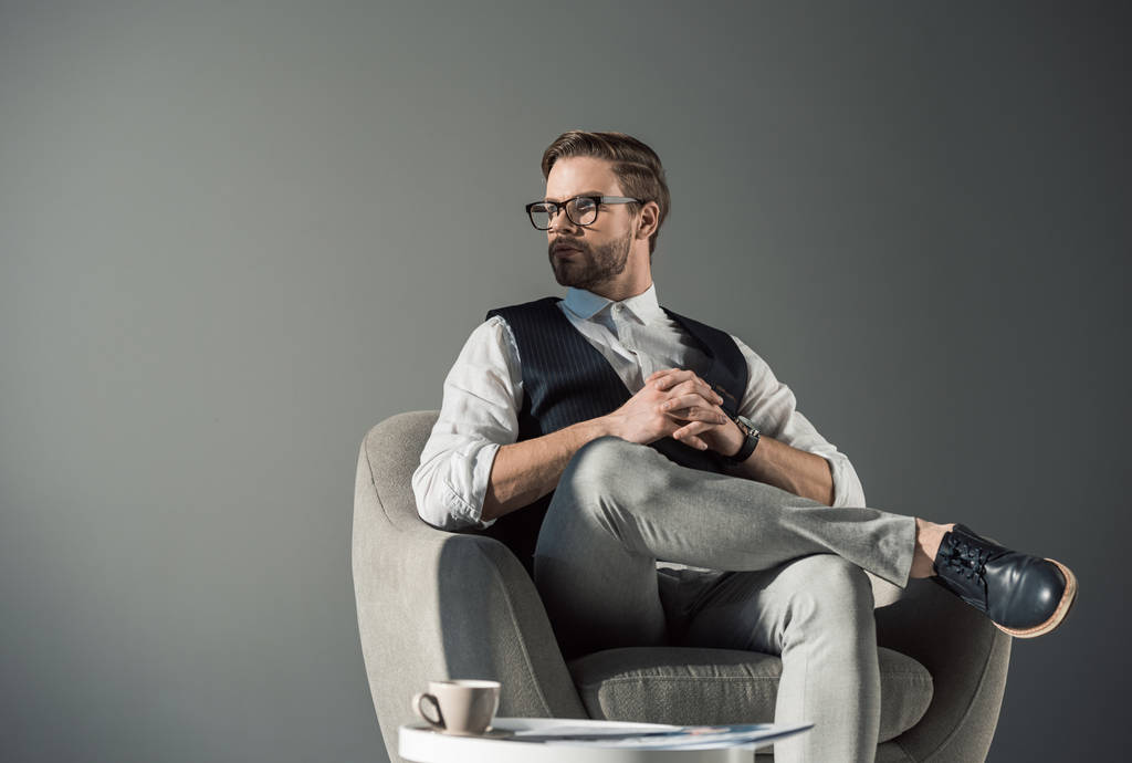 ritratto di un bel giovane uomo d'affari con gli occhiali che distoglie lo sguardo mentre è seduto in poltrona isolato sul grigio
 - Foto, immagini