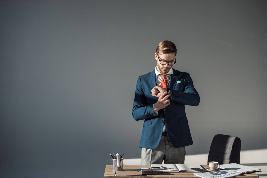 Portret van knappe jonge zakenman controleren horloge op werkplek op grijs - Foto, afbeelding