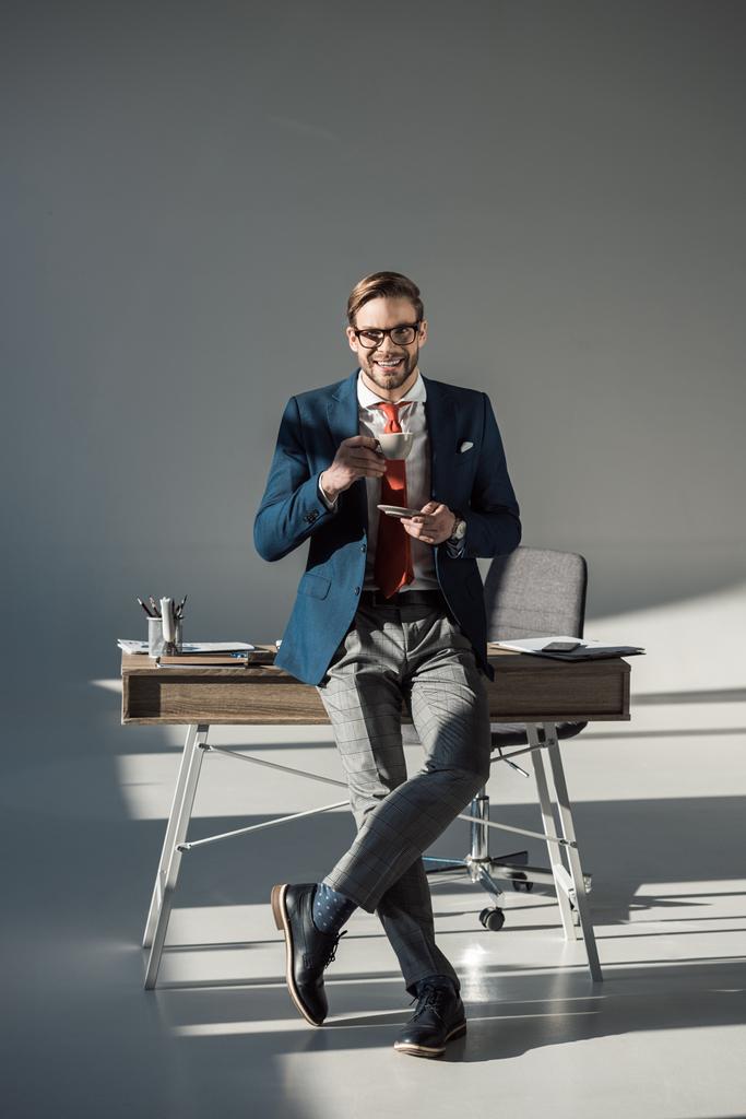 lachende stijlvolle zakenman met kop koffie zittend op tafel en kijken naar camera op grijs  - Foto, afbeelding