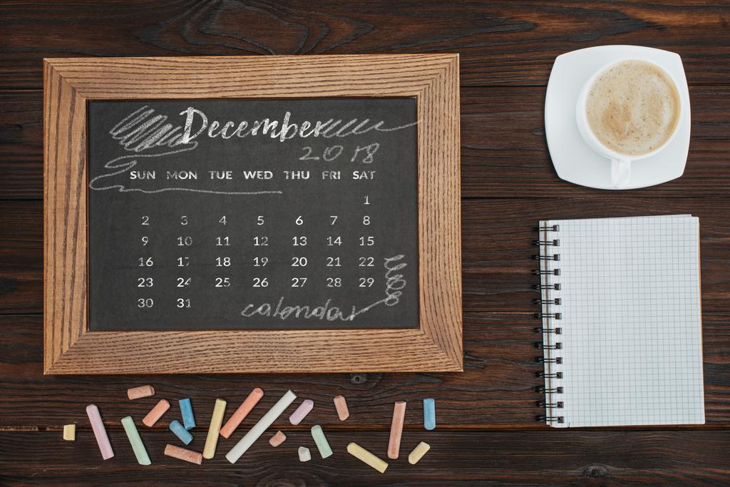 Draufsicht auf arrangierte Tasse Kaffee, leeres Notizbuch, bunte Kreiden und Tafel mit Dezemberkalender - Foto, Bild
