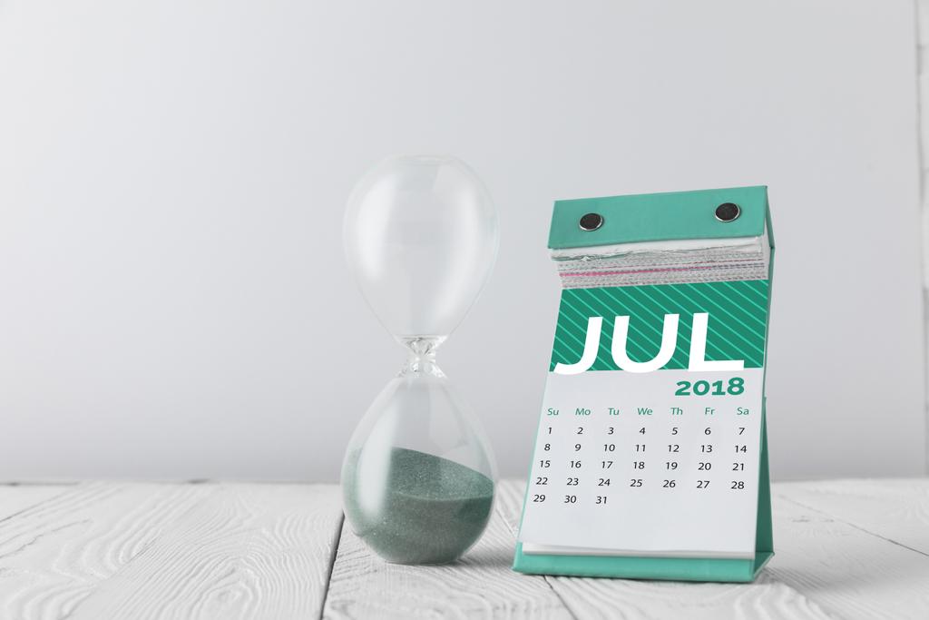 κοντινό πλάνο προβολής κλεψύδρα και ημερολόγιο Ιουλίου σε ξύλινη επιτραπέζια απομονωθεί σε λευκό - Φωτογραφία, εικόνα