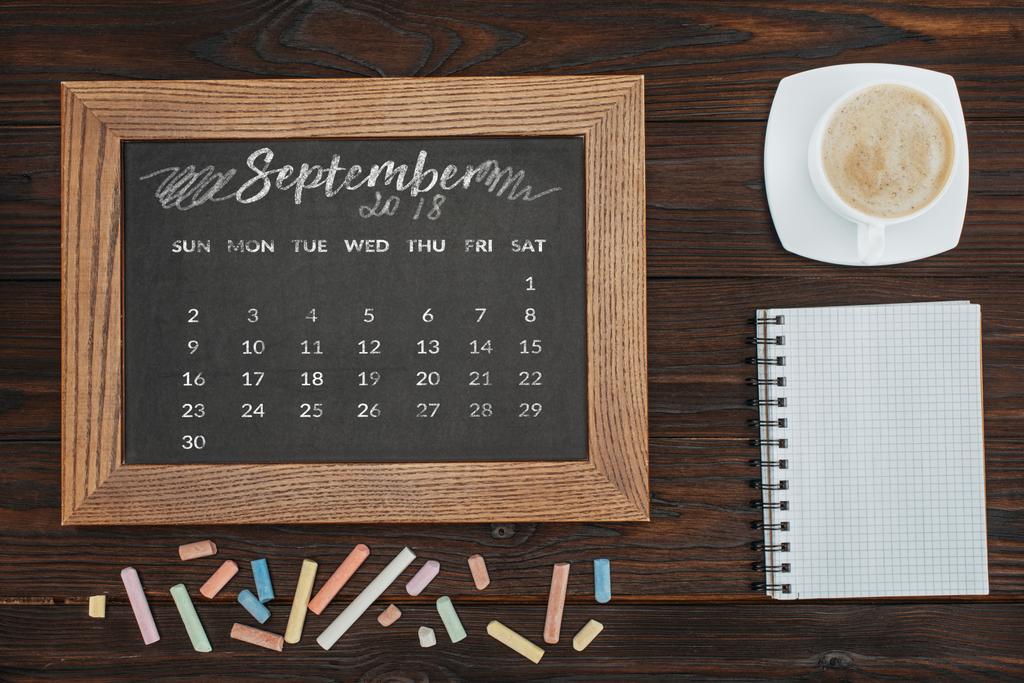 το Top view τοποθετημένα φλιτζάνι καφέ, κενό σημειωματάριο, πολύχρωμες κιμωλίες και Μαυροπίνακας με ημερολόγιο του Σεπτεμβρίου - Φωτογραφία, εικόνα