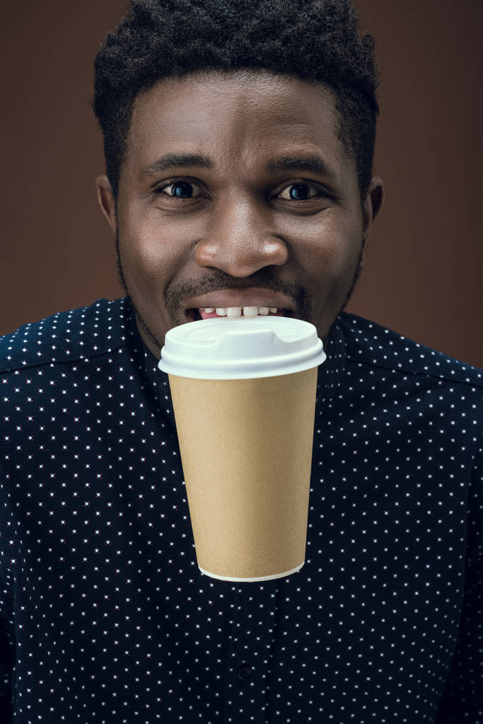 african american man holding disposable coffee cup with teeth isolated on brown - Photo, Image