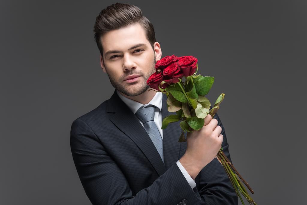 όμορφος άντρας κρατάει μπουκέτο από κόκκινα τριαντάφυλλα, απομονώνονται σε γκρι κοστούμι - Φωτογραφία, εικόνα