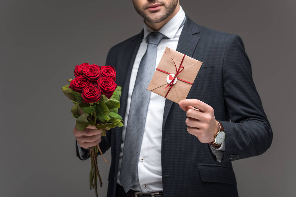 abgeschnittene Ansicht eines Mannes im Anzug mit roten Rosen und Umschlag zum Valentinstag, isoliert auf grau - Foto, Bild