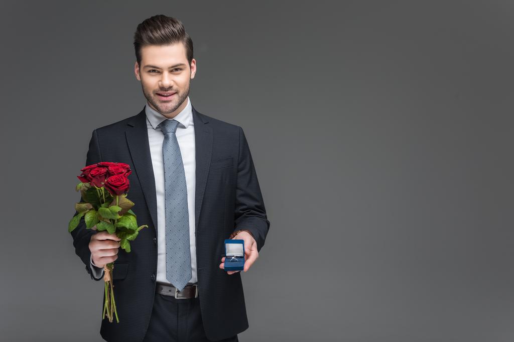 przystojny elegancki mężczyzna trzyma bukiet czerwonych róż i wniosek pierścień, na szarym tle - Zdjęcie, obraz