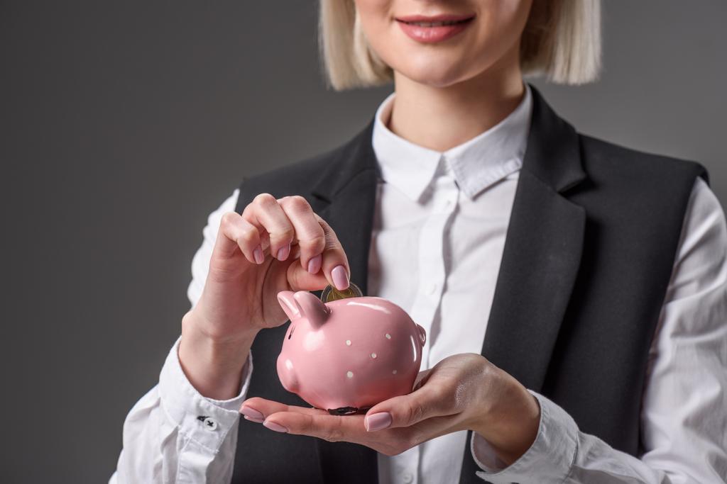 обрезанный снимок бизнесвумен положить монету в копилку поросёнка изолированы на серый
 - Фото, изображение