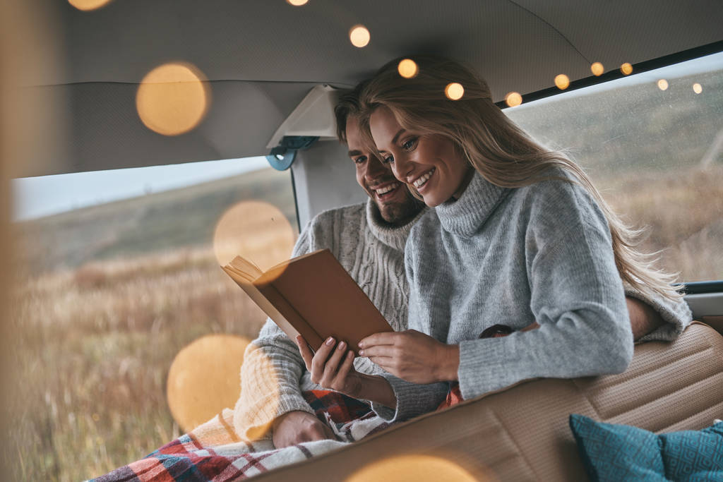 Красива молода пара читає книгу і посміхається, сидячи в стилі ретро міні фургон
 - Фото, зображення