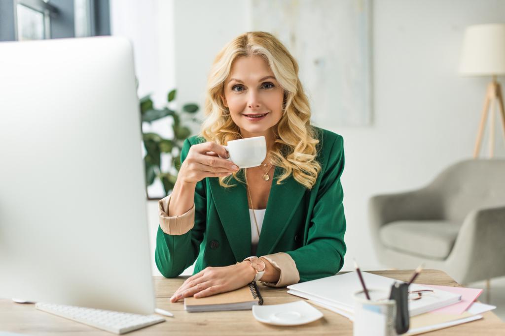 lächelnde Geschäftsfrau mittleren Alters, die Kaffee trinkt und am Arbeitsplatz einen Desktop-Computer benutzt - Foto, Bild