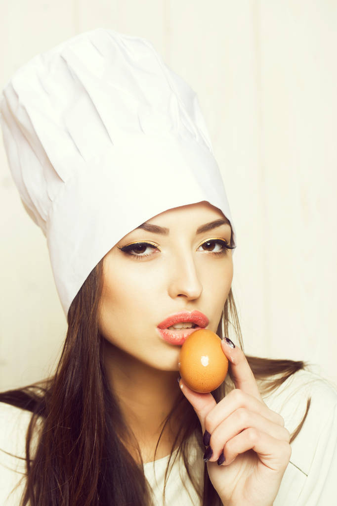 One beautiful sensual young female cook in professional white uniform of hat and apron cooking homemade food holding egg near face in kitchen on wooden background, vertical picture - Photo, Image