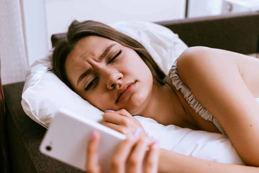 sad sleepy girl lies in bed, wakes up early, turns off alarm clock on smartphone - Photo, Image