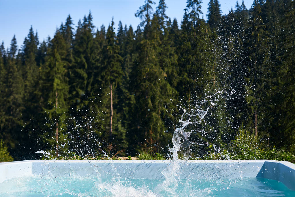Pool with splashes in air on background of coniferous forest. Place for swimming outdoors with picturesque view. Holiday, vacation, weekend, entertainment concept - Photo, Image
