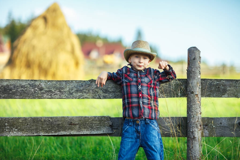 Poika nojasi selkäänsä puuaidalle, katsoi kameraan ja hymyili. Ulkomuotokuva läheltä. Pieni cowboy maatilalla poseeraa taustalla hämärä suuri heinäsuopa ja kylä maisema
 - Valokuva, kuva