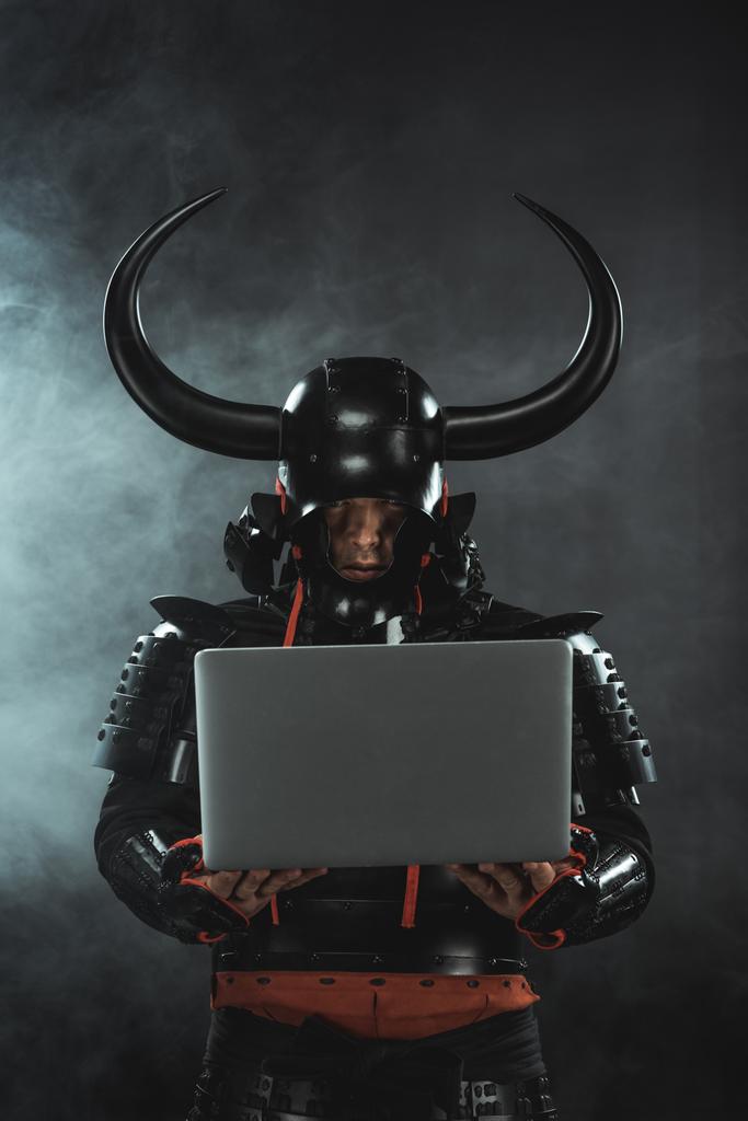 gepantserde samurai krijger met behulp van de laptop op een donkere achtergrond met rook - Foto, afbeelding