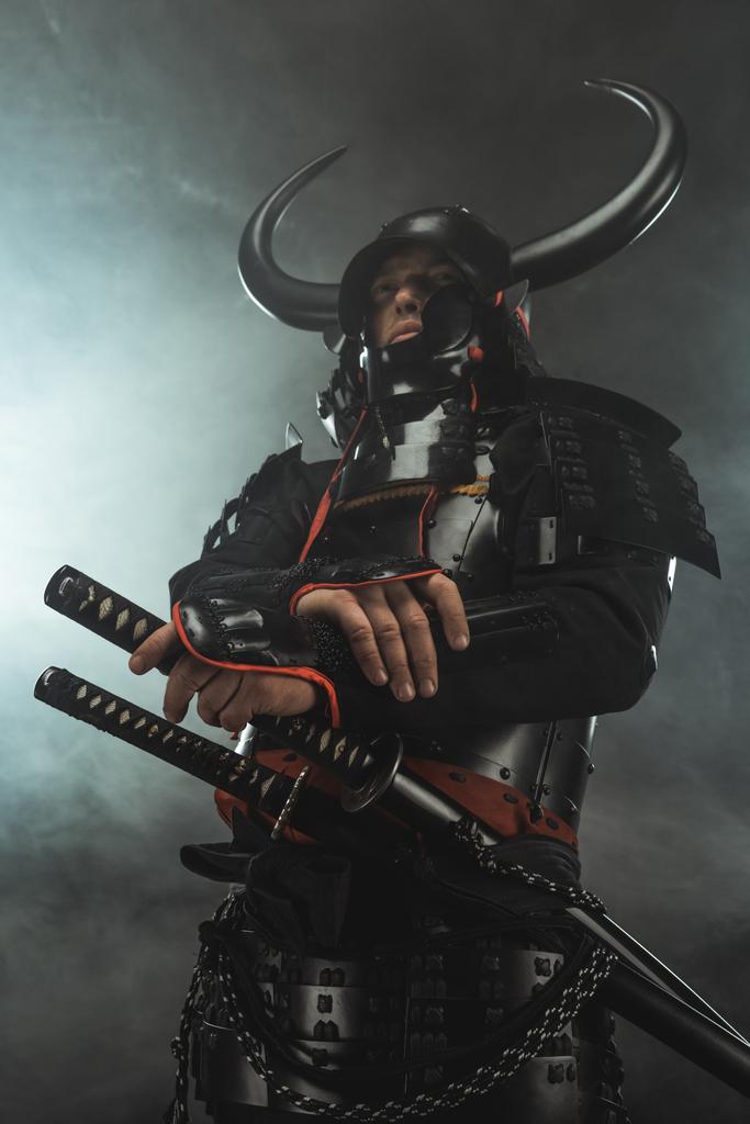 bottom view of samurai in traditional armor with swords on dark background with smoke - Photo, Image