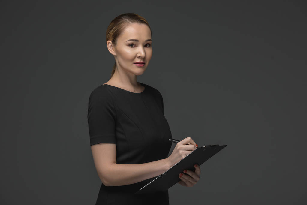 smiling kazakh businesswoman writing on clipboard and looking at camera isolated on grey - Photo, Image