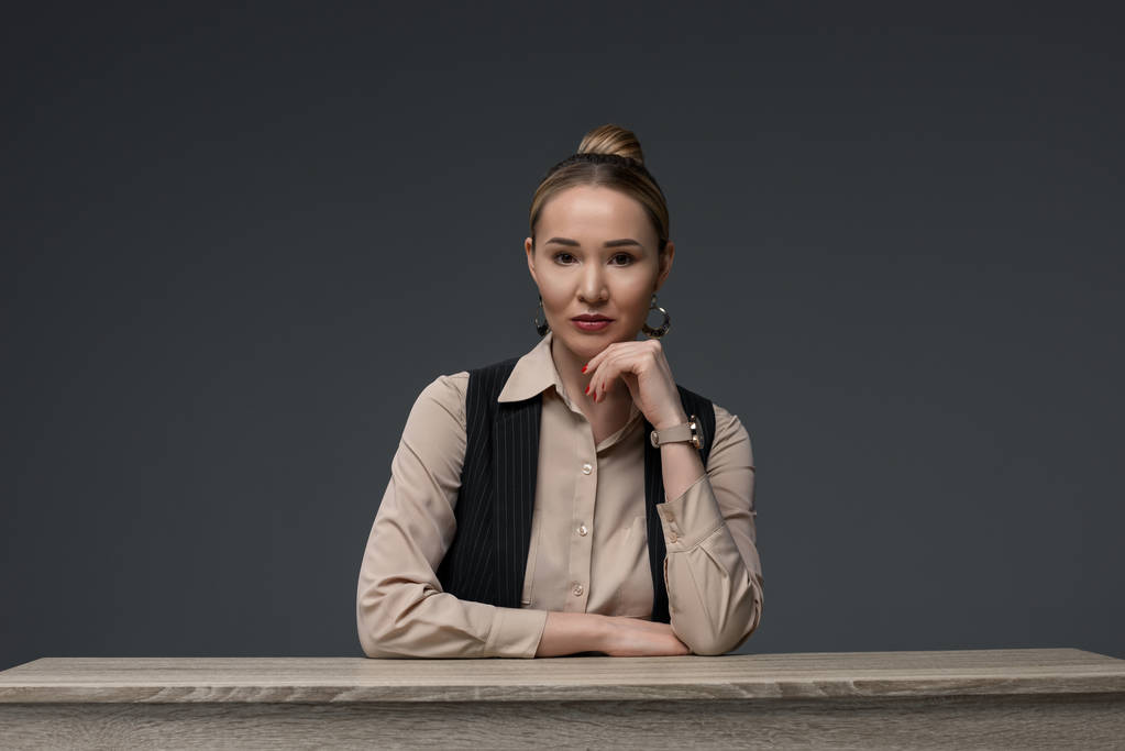 красива казахська бізнес-леді сидить за столом і дивиться на камеру ізольовано на сірому
 - Фото, зображення