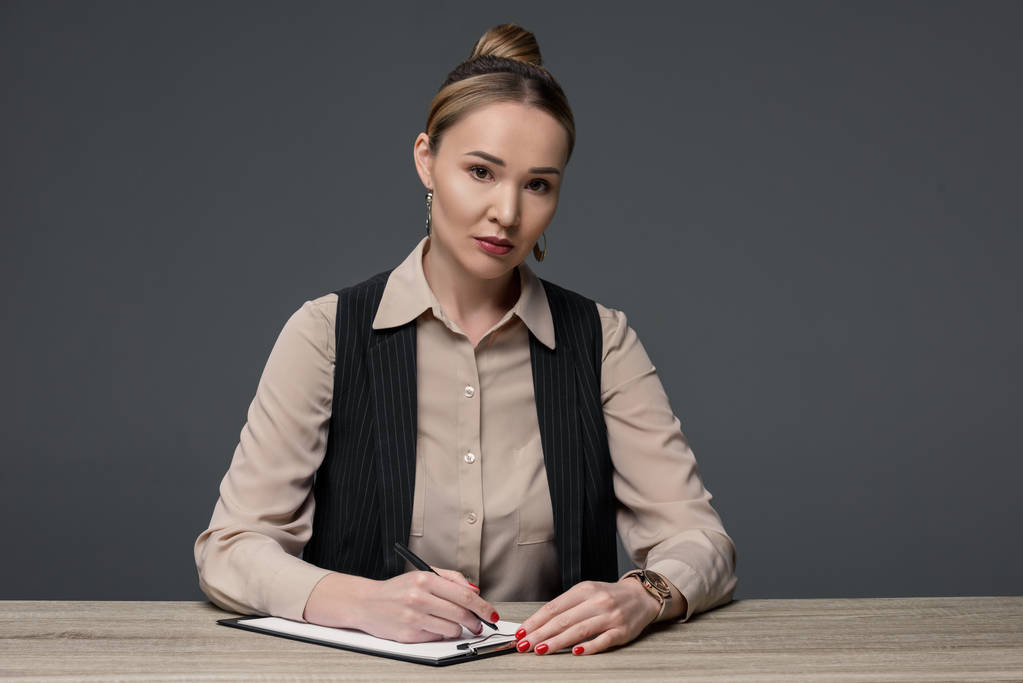 kazakh businesswoman writing on clipboard and looking at camera isolated on grey - Photo, Image