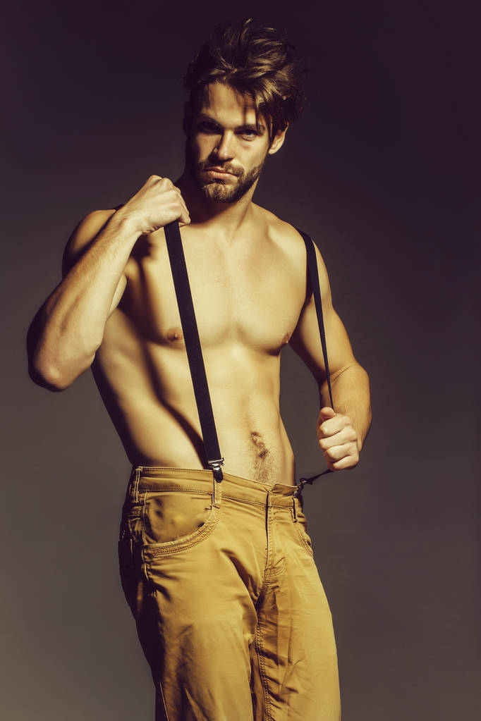 Handsome guy fashion sexy young bearded macho man model with suspenders on pants has bare muscular torso holds on grey background - Photo, Image