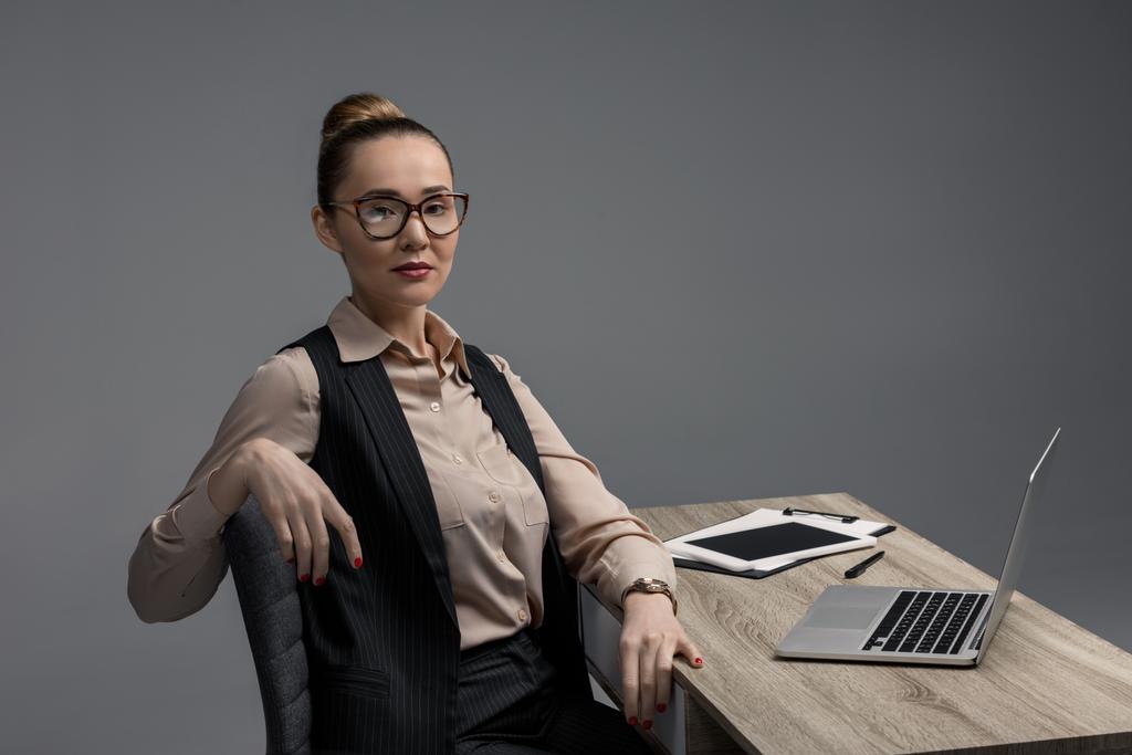 attractive kazakh businesswoman in eyeglasses using laptop and looking at camera isolated on grey - Photo, Image
