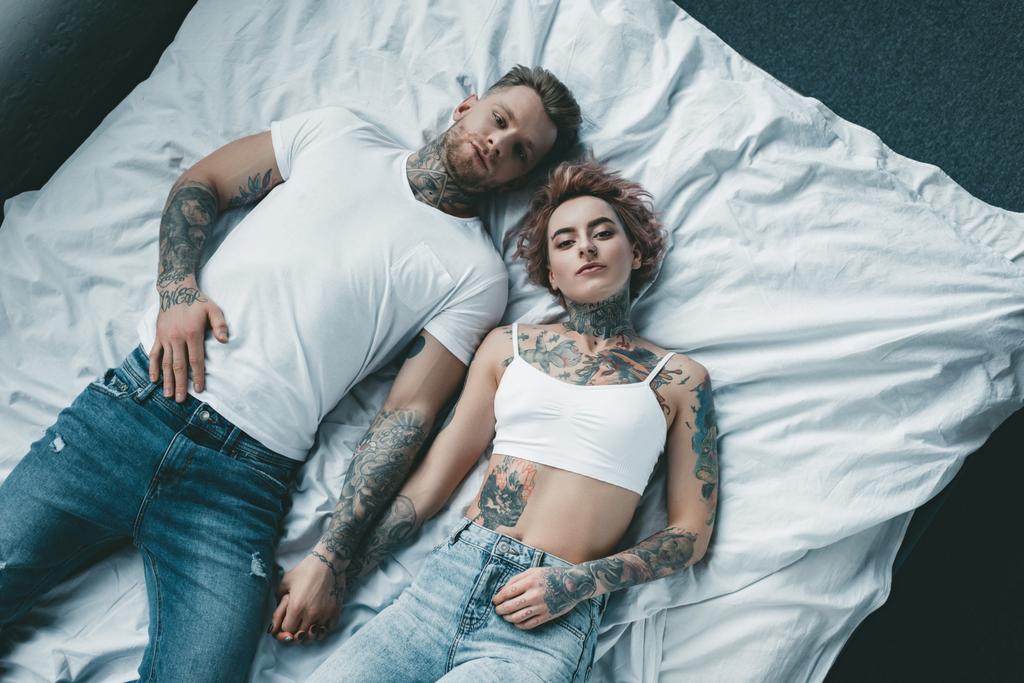 Draufsicht auf tätowiertes junges Paar, das Hände hält und auf dem Bett liegt - Foto, Bild