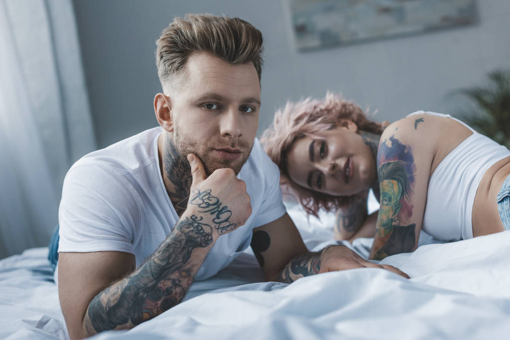 όμορφο τατουάζ ζευγάρι βλέπει κάμερα ενώ χαλαρώνετε στο κρεβάτι  - Φωτογραφία, εικόνα