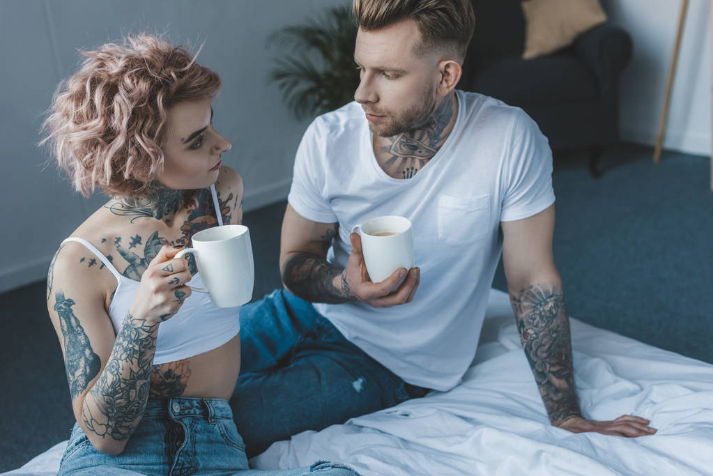 jovem casal tatuado olhando um para o outro e beber café na cama de manhã
 - Foto, Imagem