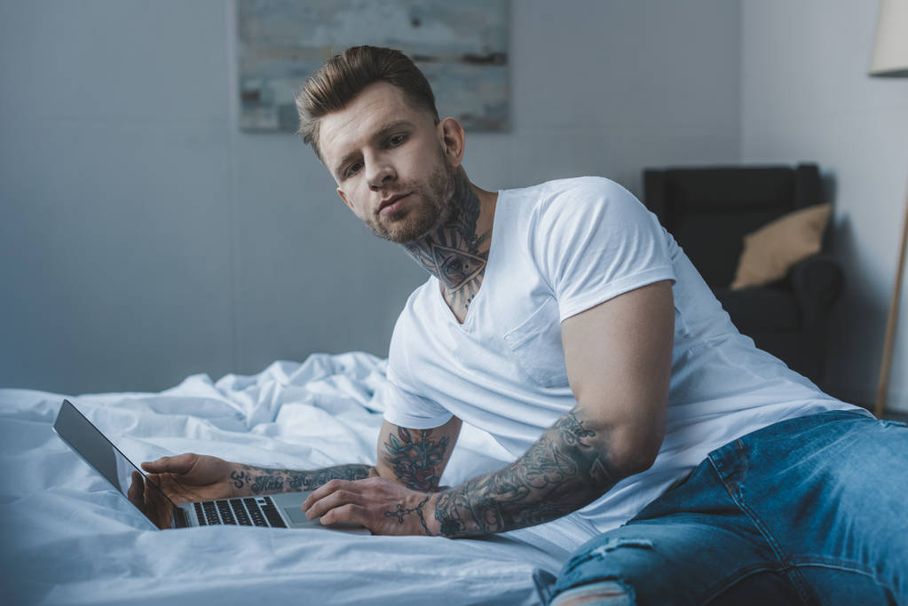 Tetovaný muž dívá na kameru při používání notebooku v posteli  - Fotografie, Obrázek