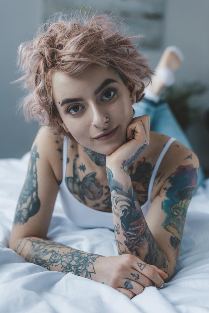 tätowiertes Mädchen mit rosa Haaren liegt auf dem Bett und blickt in die Kamera - Foto, Bild