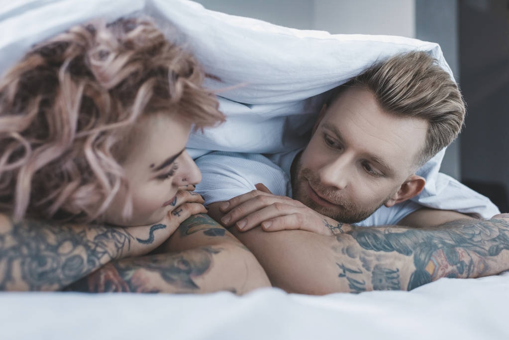 молодая татуированная пара смотрит друг на друга под белым одеялом в спальне
 - Фото, изображение