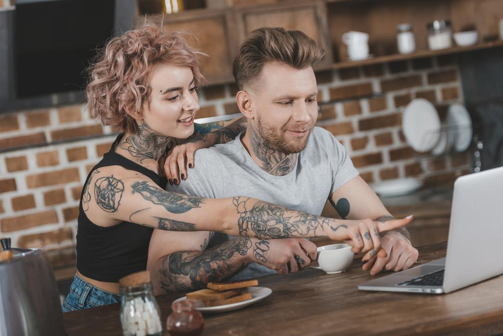 coppia tatuata che punta al computer portatile durante la colazione in cucina
 - Foto, immagini