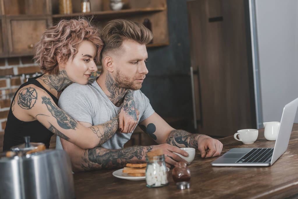 tattooed couple hugging and using laptop during breakfast in kitchen - Photo, Image
