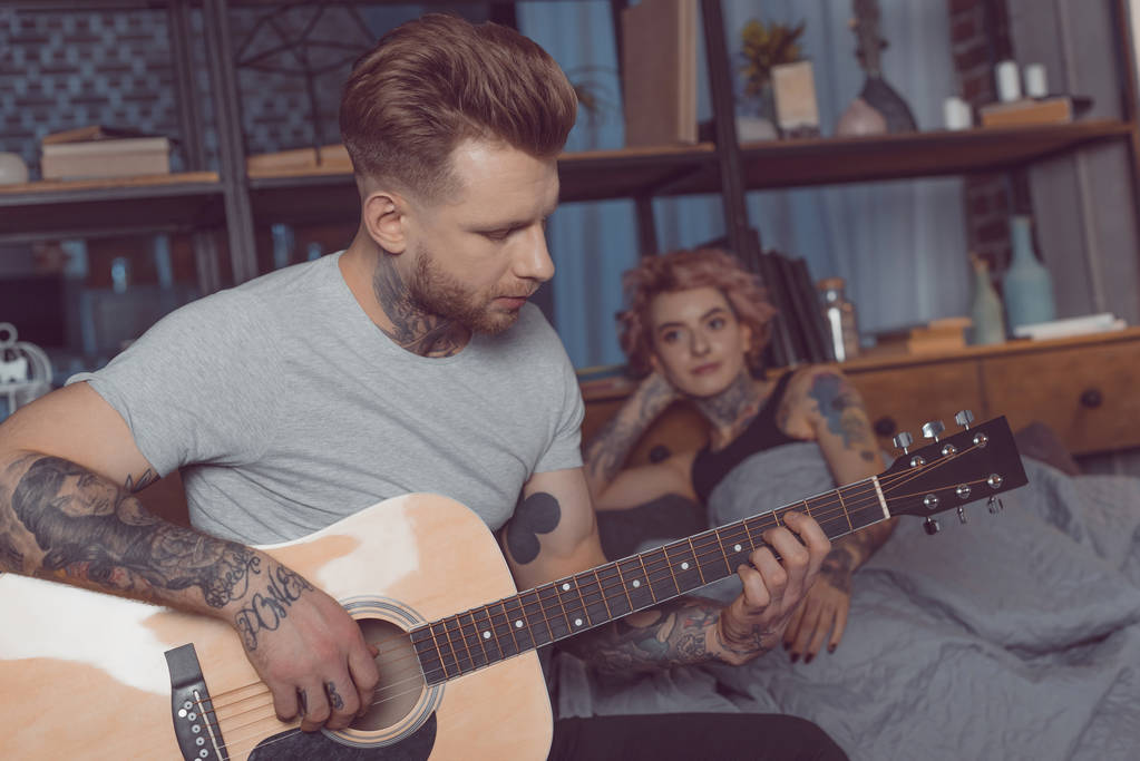 tetovált barátja, a barátnője otthon játszik akusztikus gitár - Fotó, kép