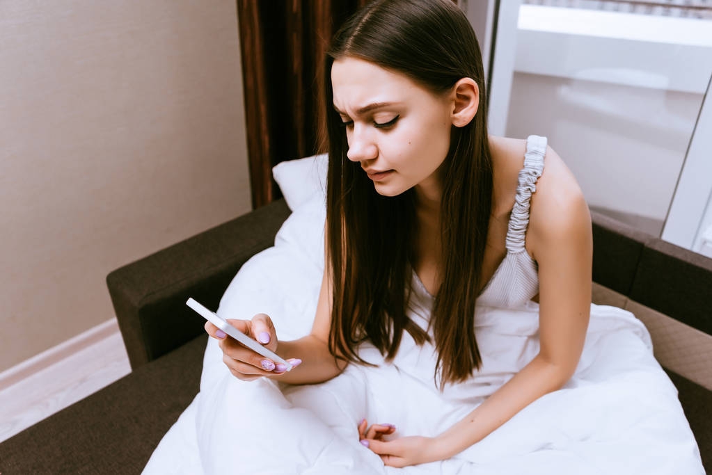 сонна молода дівчина сидить у ліжку під білою ковдрою, дивиться в її смартфон
 - Фото, зображення
