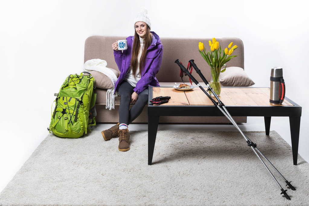 junge Frau in warmer Kleidung mit Rucksack und Wanderausrüstung trinkt zu Hause Tee - Foto, Bild