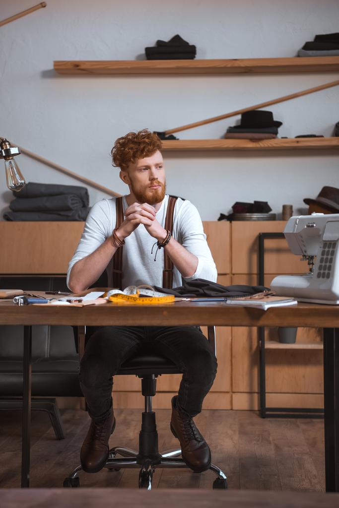 pensativo joven diseñador de moda masculina mirando hacia otro lado mientras está sentado en el lugar de trabajo
 - Foto, Imagen