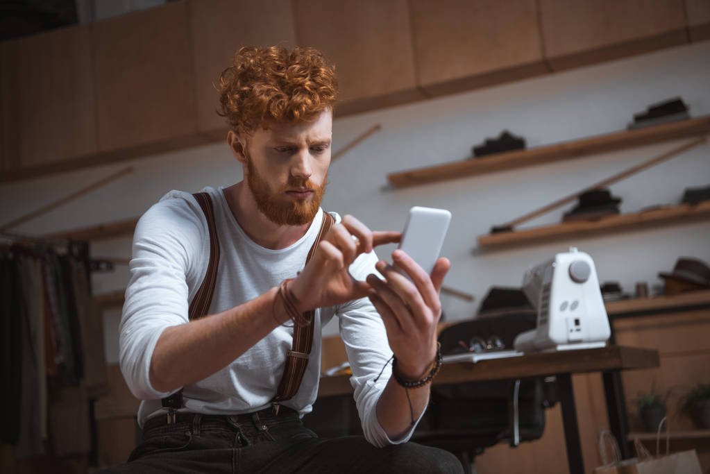 enfocado joven diseñador de moda masculino usando teléfono inteligente mientras está sentado en el taller
 - Foto, imagen