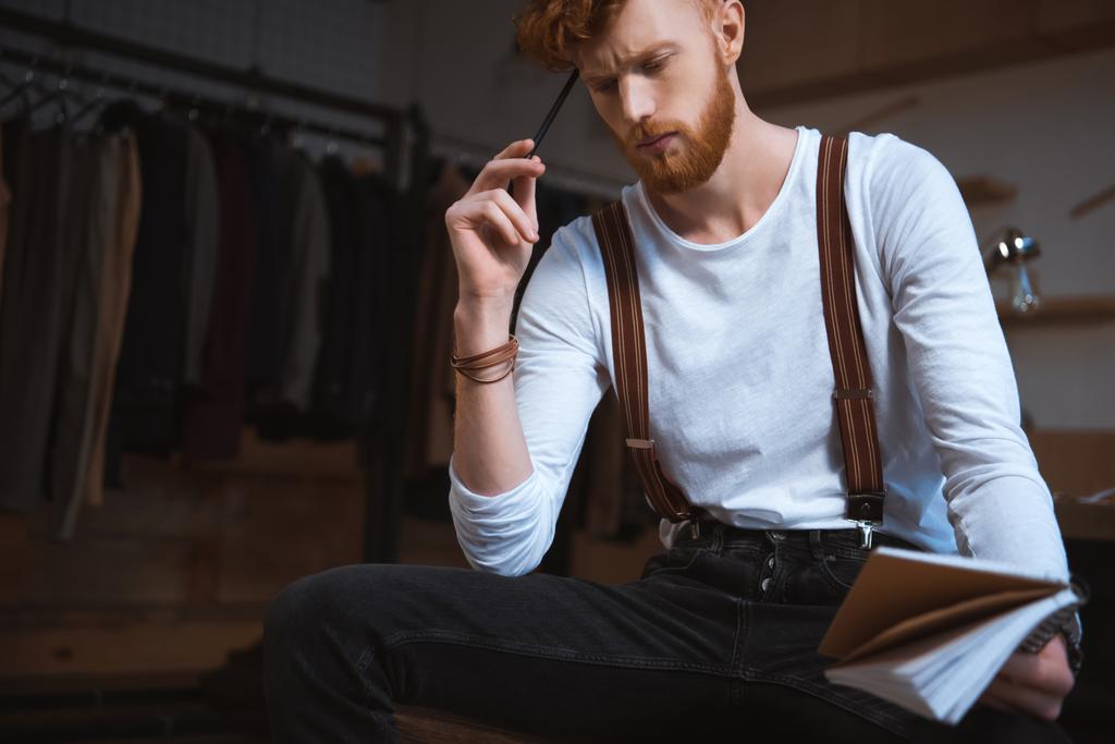 pensativo jovem designer de moda masculina com notebook e lápis sentado no local de trabalho
 - Foto, Imagem