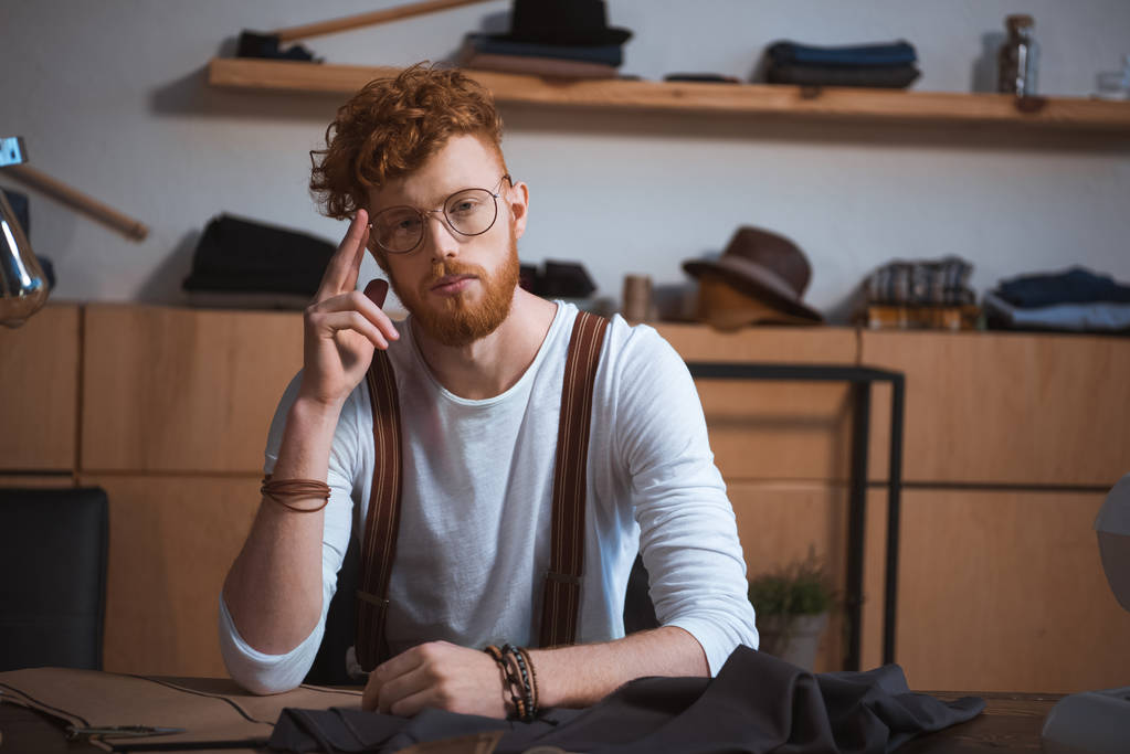 przystojny młody projektant mody w okularach, patrząc na kamery, siedząc w miejscu pracy - Zdjęcie, obraz