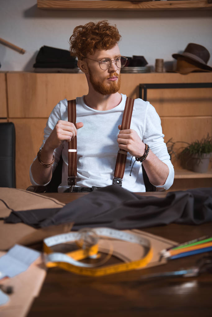 zamyšlený mladý vousatý módní návrhář v uhýbaje zatímco sedí na pracovišti brýlích - Fotografie, Obrázek