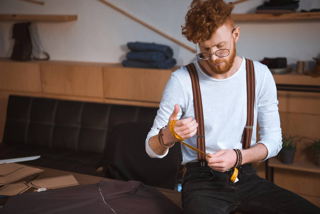 молодий дизайнер чоловічої моди в окулярах тримає вимірювальну стрічку в майстерні
 - Фото, зображення